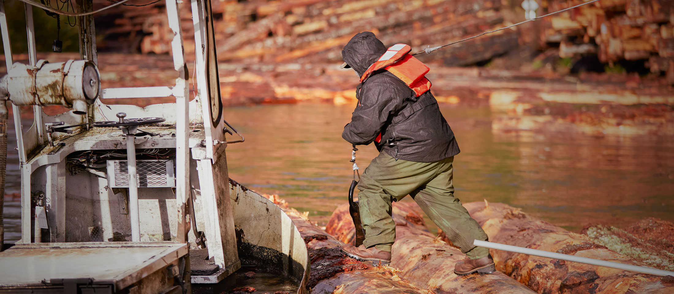 A man wearing the Valdor 8" Waterproof Comp Toe Insulated Logger boots in brown and carrying a fishing pole and walking along logs to get to their boat.